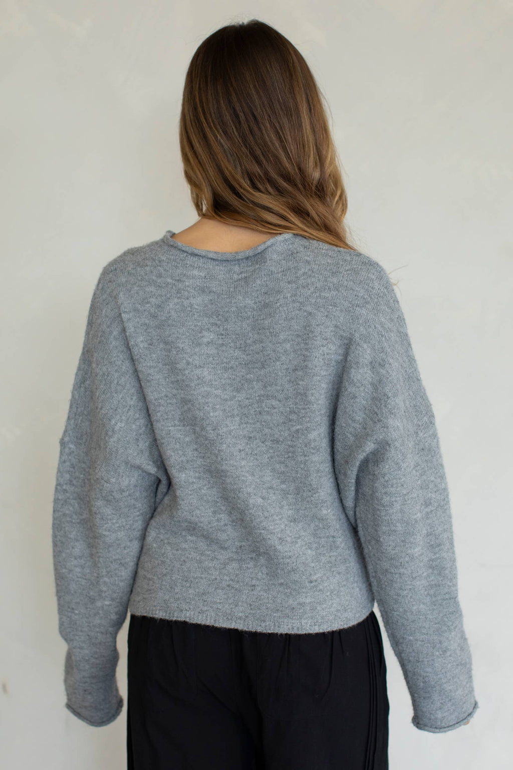 A female model facing backwards wearing a light gray cardigan, paired with black denim jeans in front of a gray studio background. 