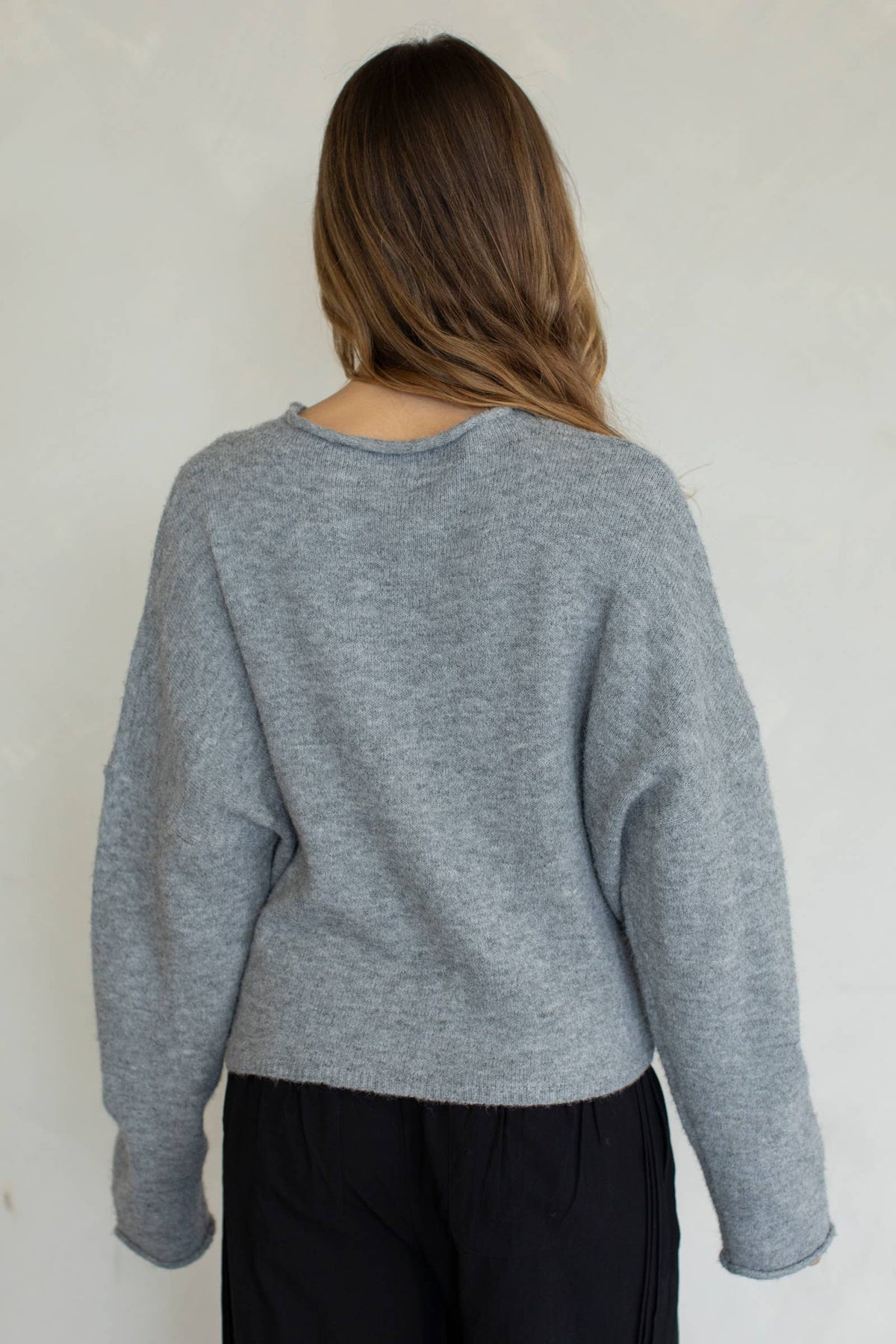 A female model facing backwards wearing a light gray cardigan, paired with black denim jeans in front of a gray studio background. 