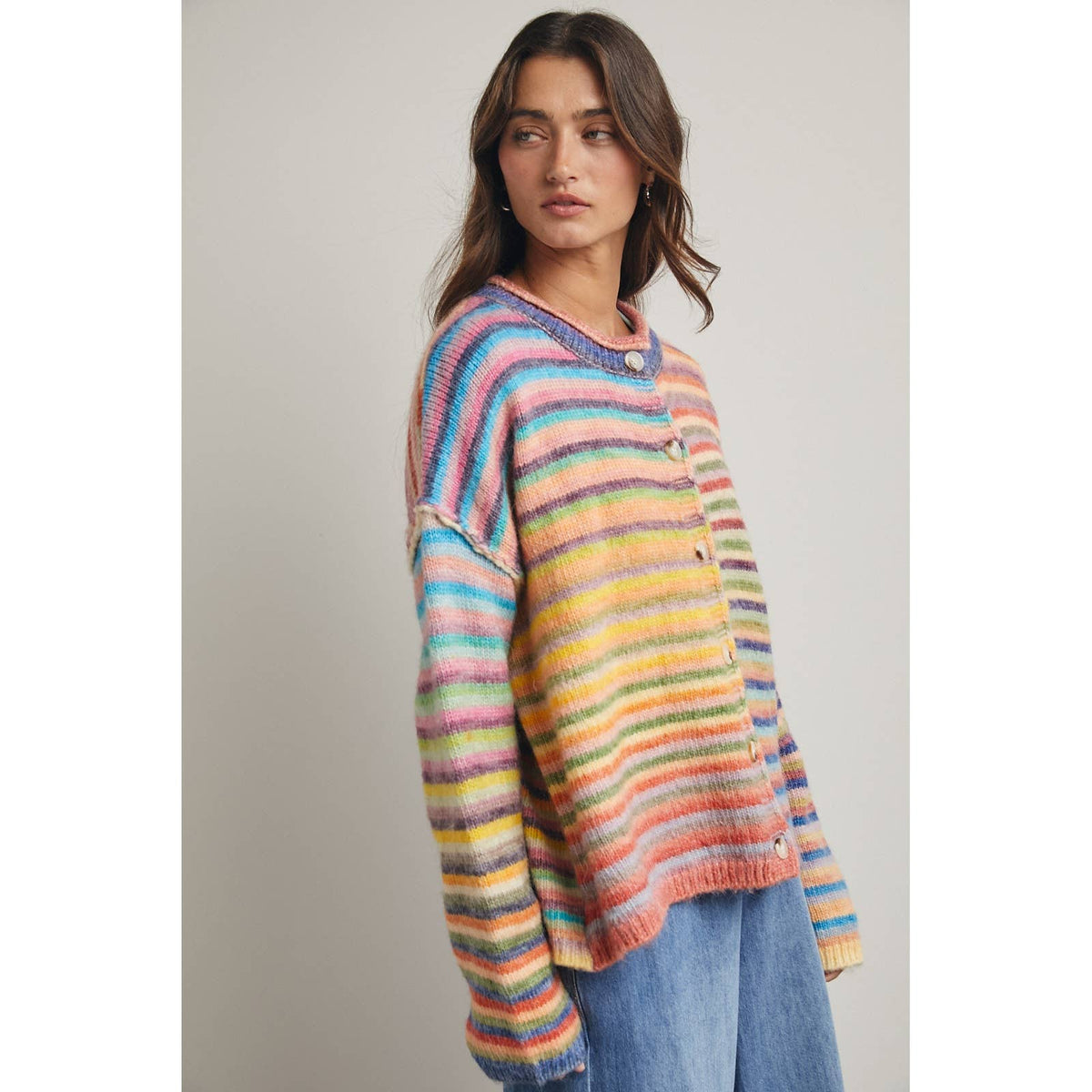 A woman wearing a colorful, rainbow fuzzy horizontal striped cardigan sweater with buttons, paired with blue jeans, against a plain light gray background.