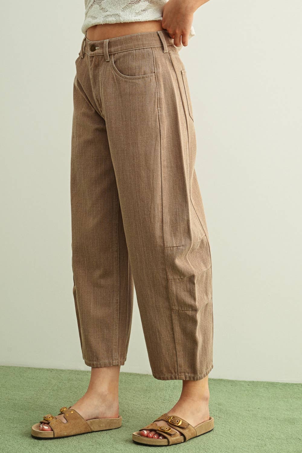 A person wearing wide-leg barrel cropped pants in a warm taupe-brown color, paired with tan double-strap sandals and a light-colored knit top, facing sideways, standing against a plain light wall with a green floor.