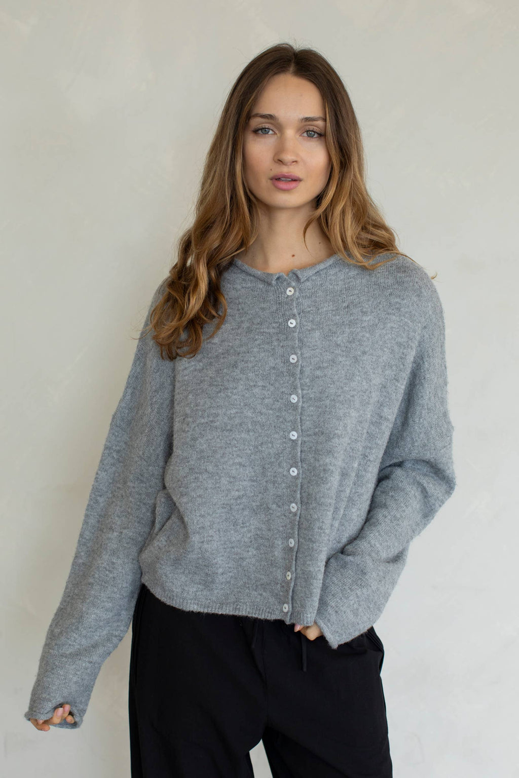 A female model wearing a light gray button up cardigan with white buttons, paired with black denim jeans in front of a gray studio background. 