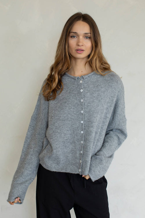 A female model wearing a light gray button up cardigan with white buttons, paired with black denim jeans in front of a gray studio background. 