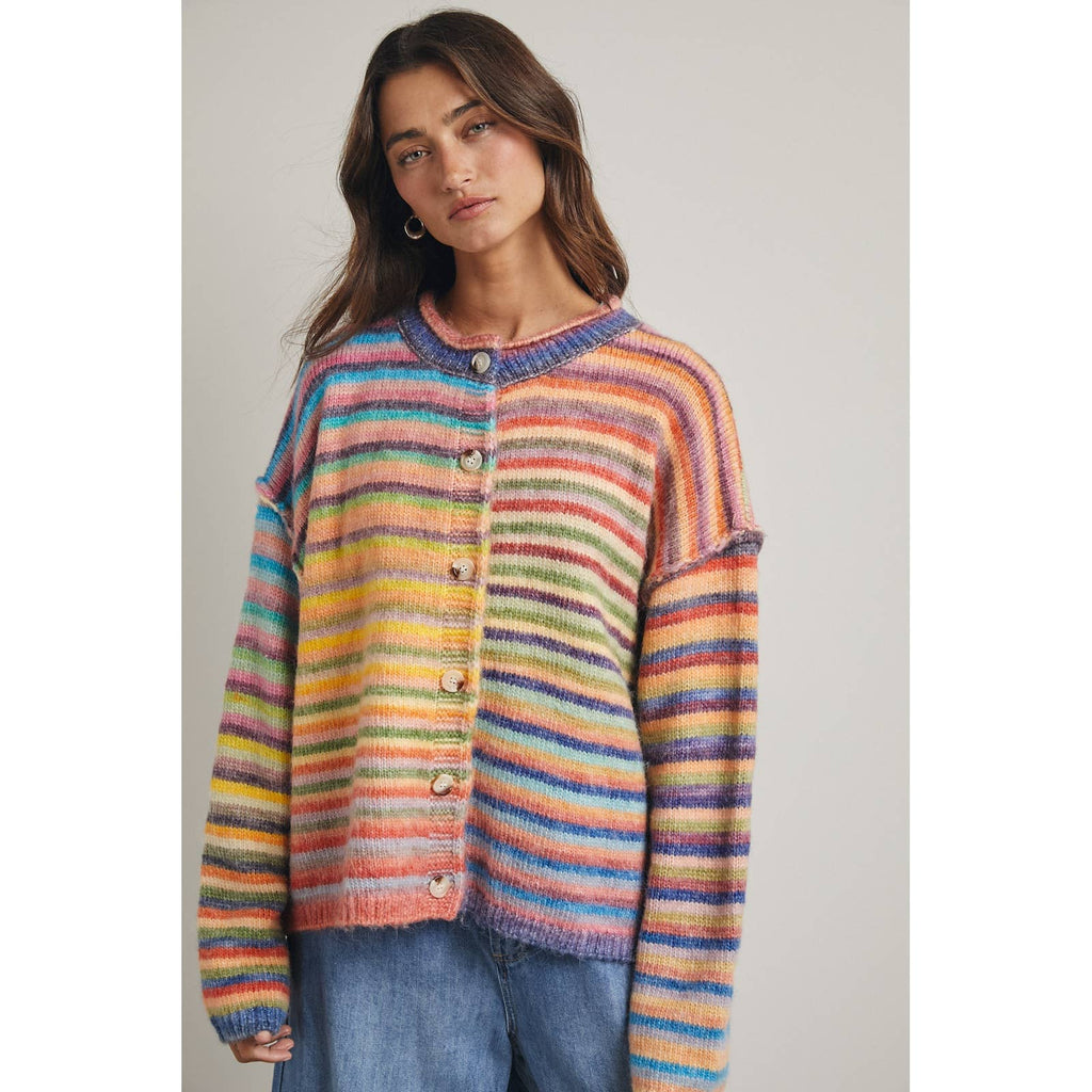 A woman wearing a colorful, rainbow fuzzy horizontal striped cardigan sweater with buttons, paired with blue jeans, against a plain light gray background.