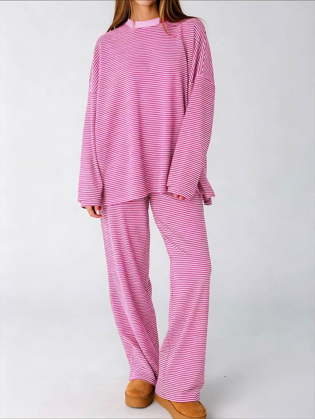 A female model wearing a pink and red striped two-piece lounge set consisting of a long-sleeve top and matching pants, paired with tan slip-on shoes, against a plain light gray background.