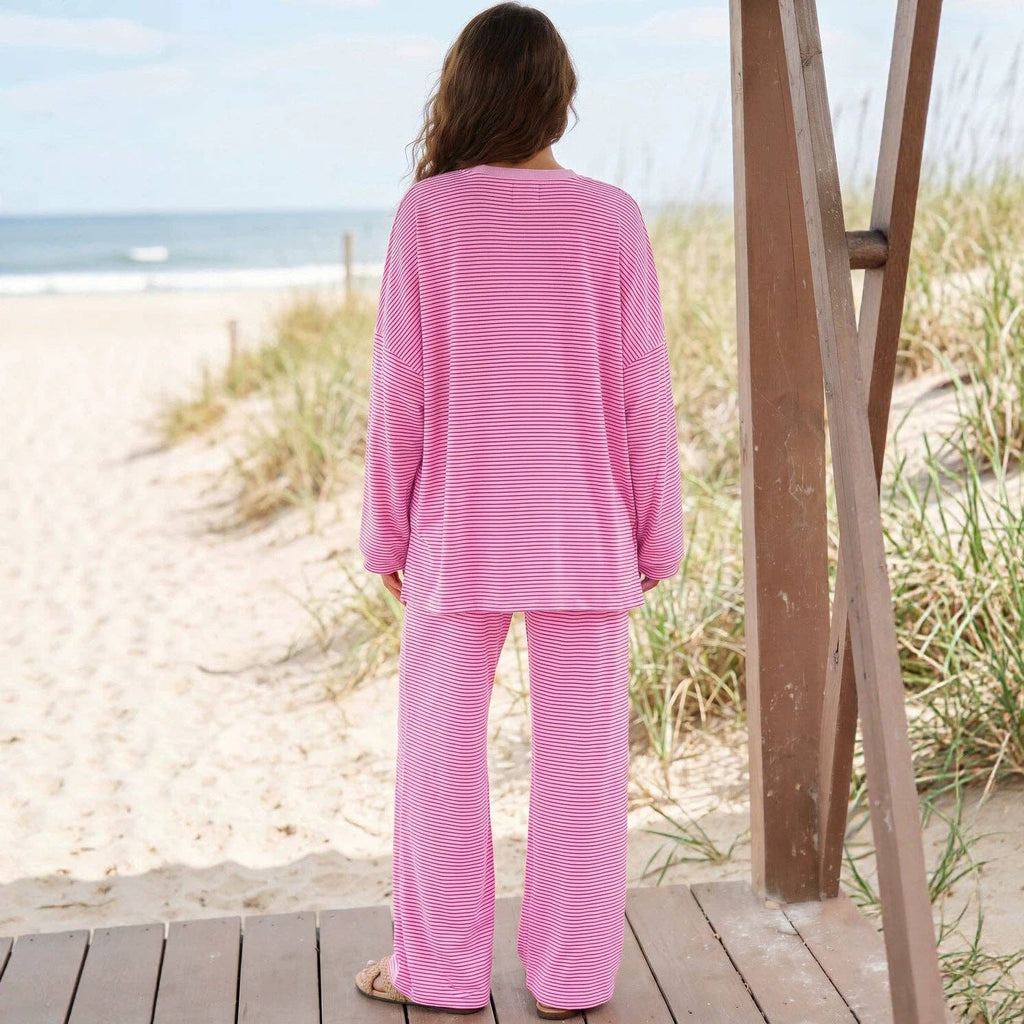A female model facing backwards wearing a pink and red striped two-piece lounge set consisting of a long-sleeve top and matching pants, paired with tan slip-on shoes, standing on a platform facing the beach. 