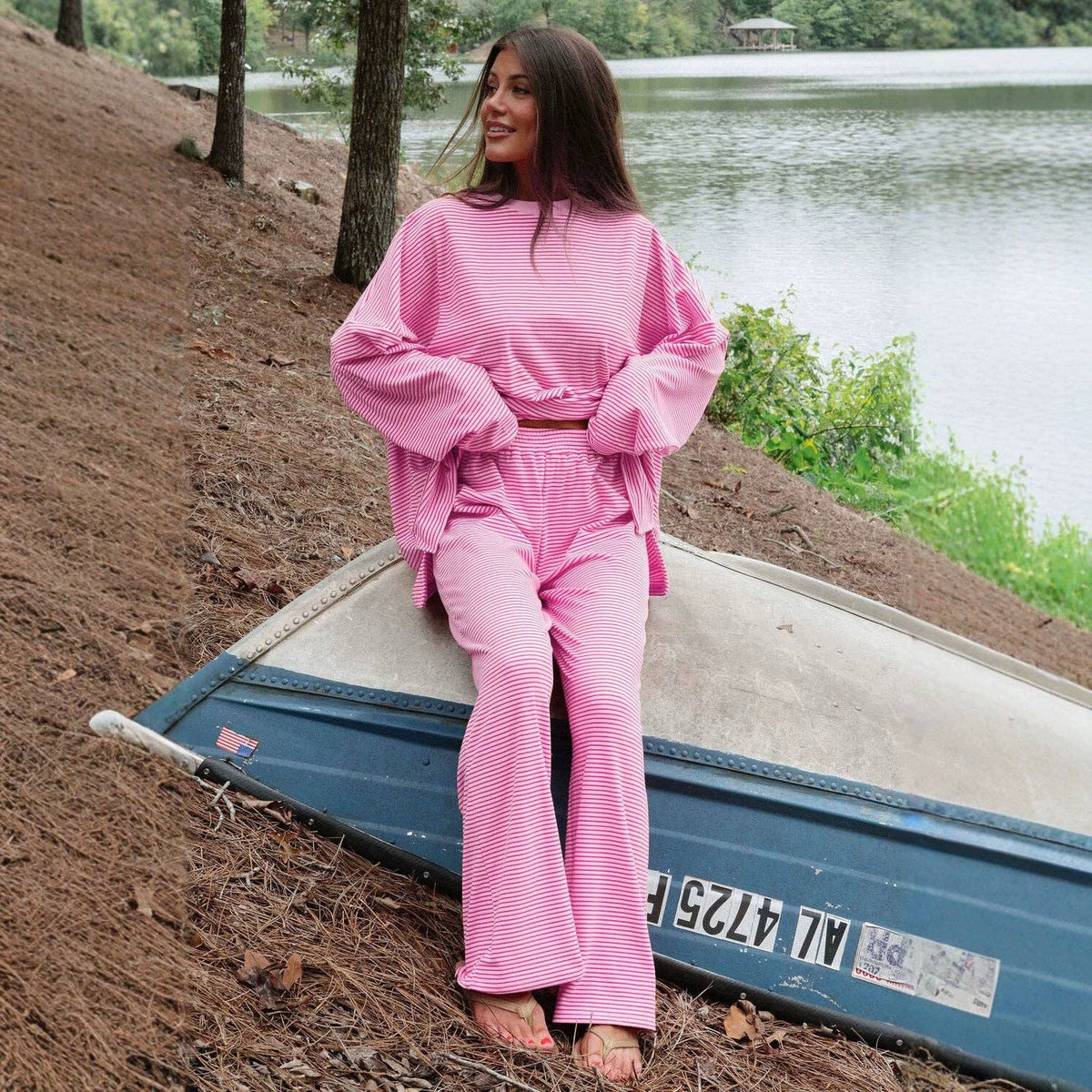 A female model wearing a pink and red striped two-piece lounge set consisting of a long-sleeve top and matching pants, paired with tan slip-on sandals. The model is sitting on an overturned boat on the bank of a lake. 