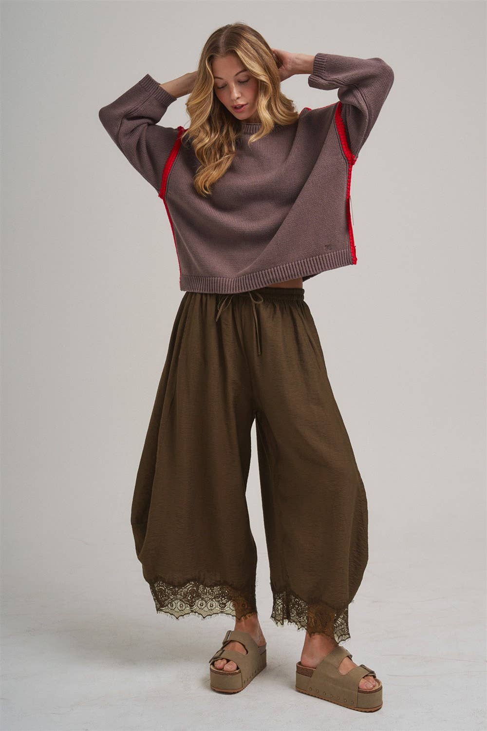 A woman modeling a cropped mauve knit sweater with red side panels and brown wide-leg pants with lace trim, paired with brown platform sandals, against a light gray studio background.