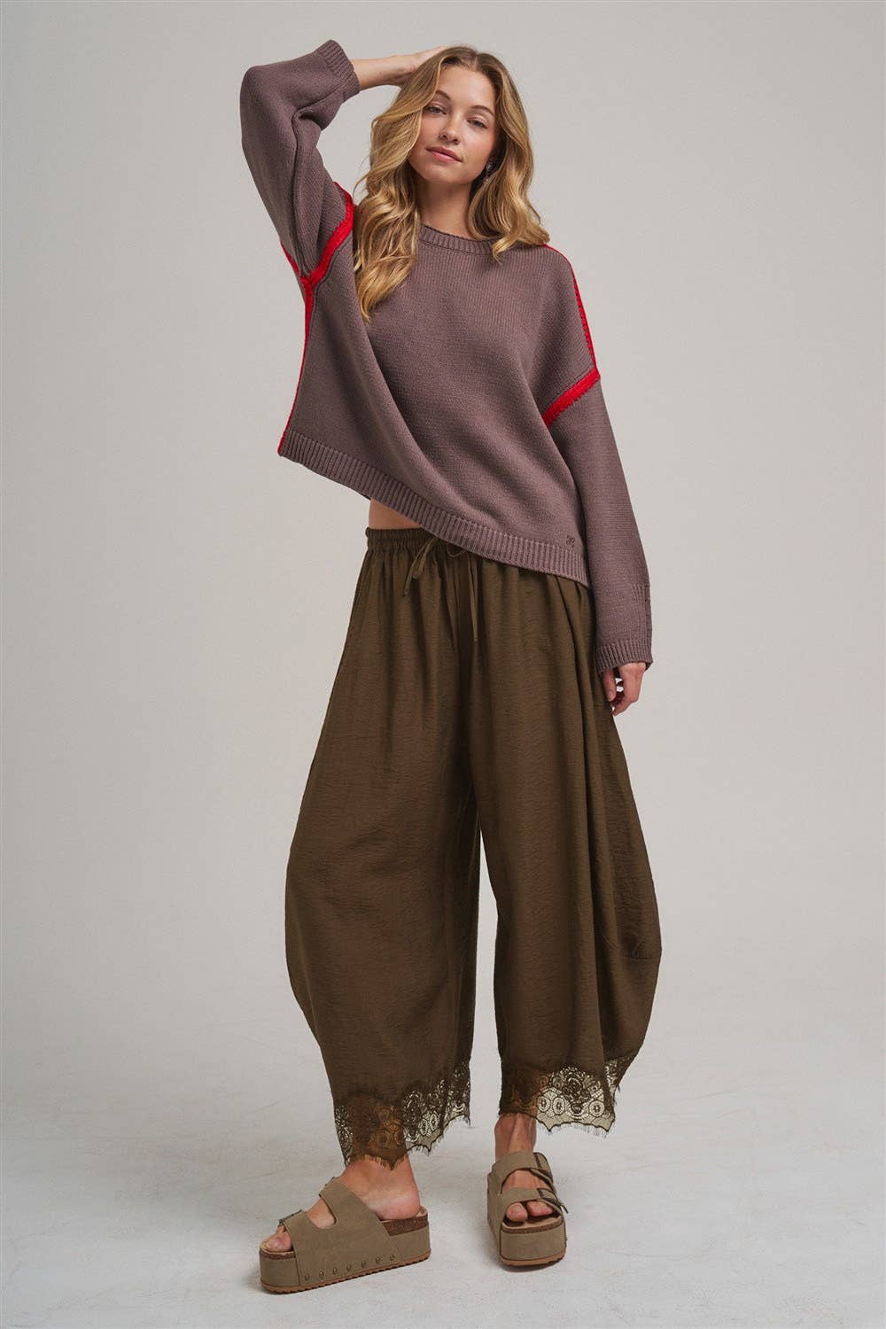 A woman modeling a cropped, oversized knit sweater in muted mauve-brown with red stripe accents on the sleeves, paired with wide-leg brown pants with lace trim and brown platform sandals, against a plain light gray studio background.